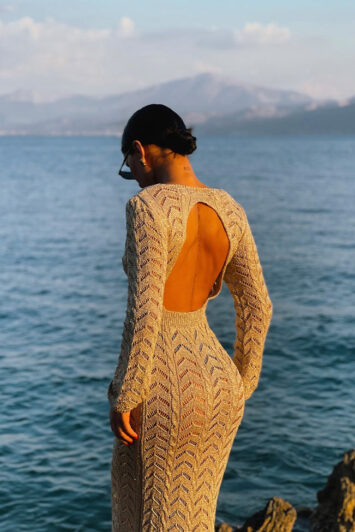 Γυναίκα με πλεκτό see-through φόρεμα σε χρυσό χρώμα ποζάρει μπροστά στη θάλασσα