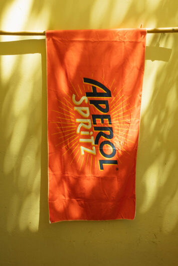 Πορτοκαλί πετσέτα παραλίας με logo Aperol Spritz κρεμασμένη σε τοίχο