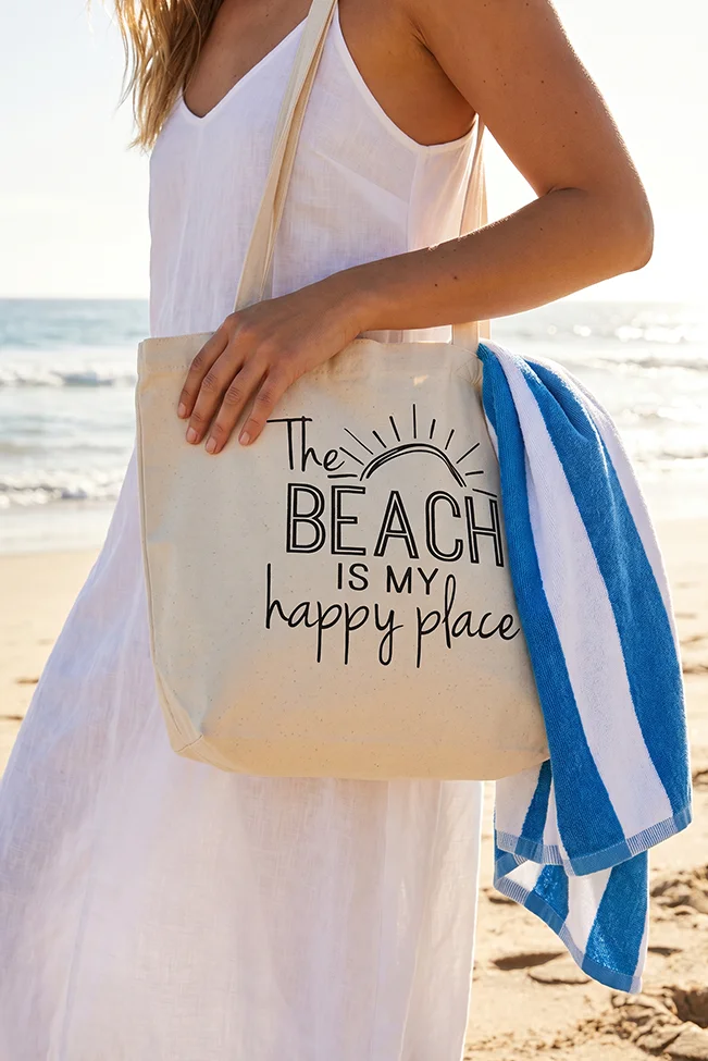 tsanta-paralias-happy-place-canvas-100181064-1-lapop-3 Γυναίκα κρατά καμβάς τσάντα παραλίας happy place στην ακρογιαλιά
